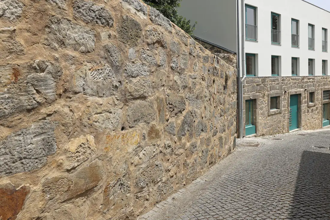 A cobblestone street runs beside a tall stone wall and a white building.