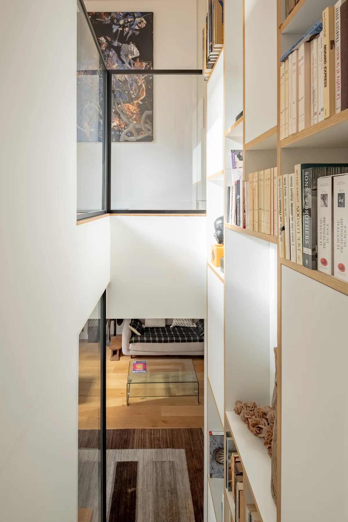 A narrow study passage has built-in bookshelves beside a glass partition overlooking a desk.