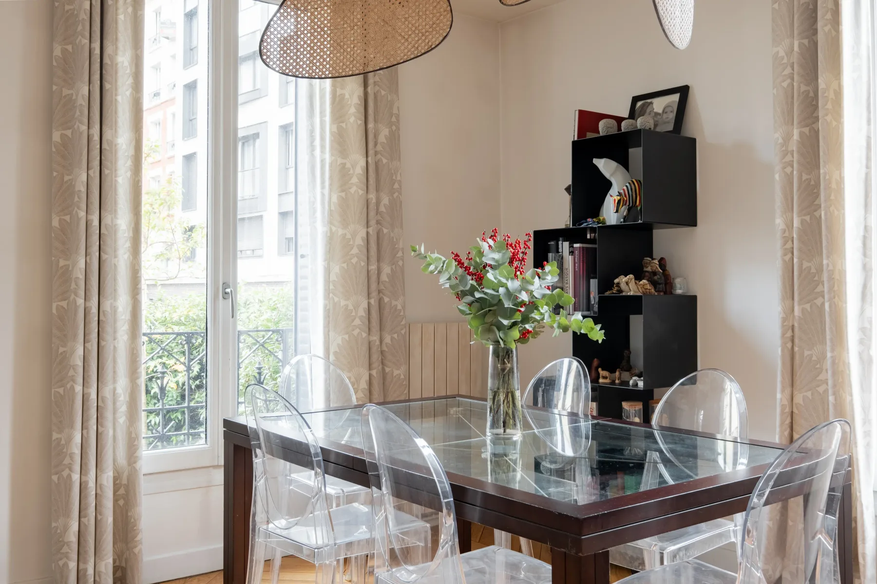 A dining area with a glass table, clear seating, and a vase of flowers by the windows.