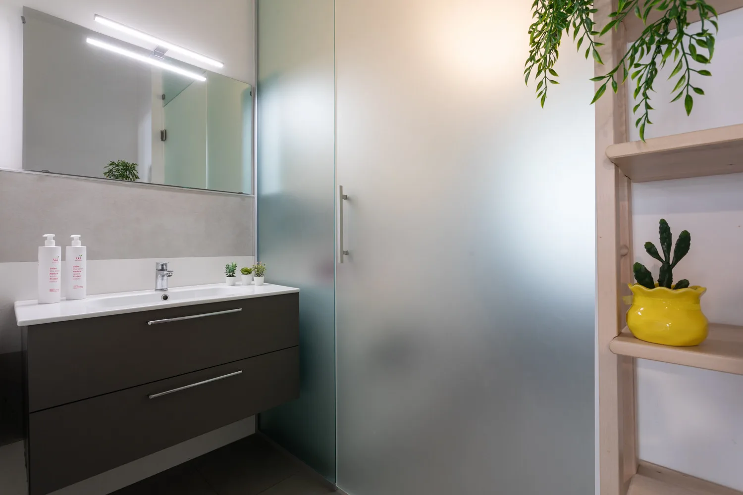 A modern bathroom vanity sits beside a frosted glass shower and a small display shelf.