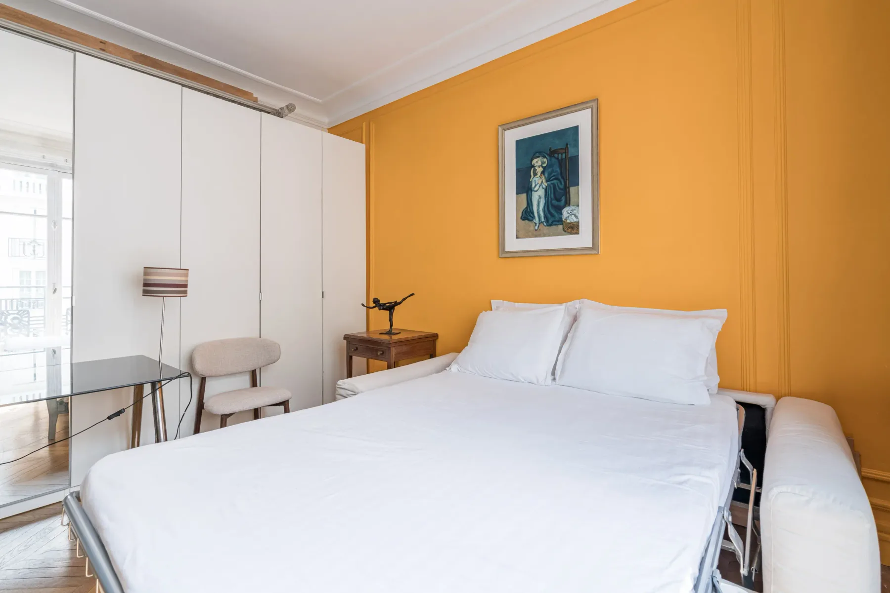 Dormitorio con una cama blanca, una pared naranja y un cuadro enmarcado.