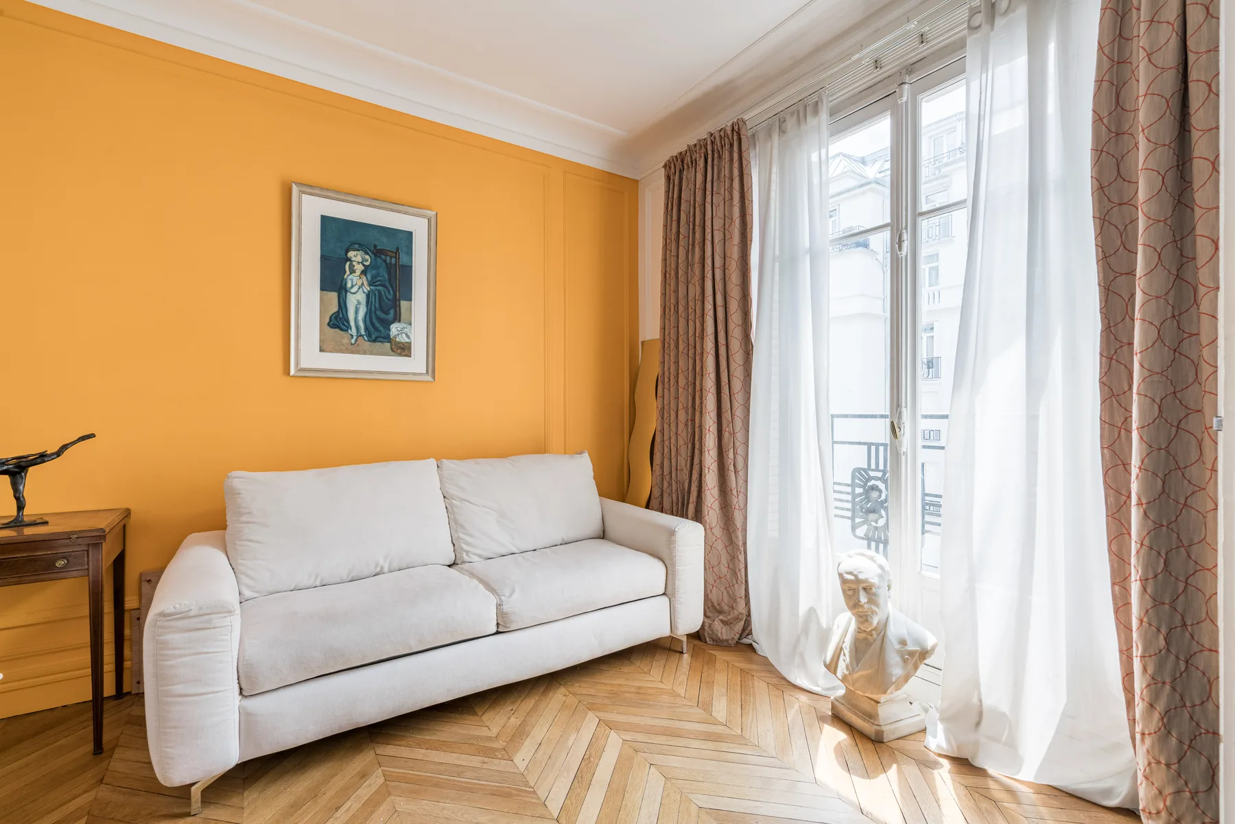 Una sala luminosa con un sofá blanco junto a ventanas altas con cortinas.