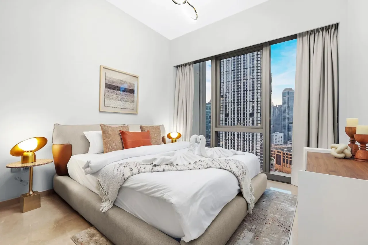 Un dormitorio luminoso con una cama tendida junto a una ventana amplia con vistas a edificios de la ciudad.