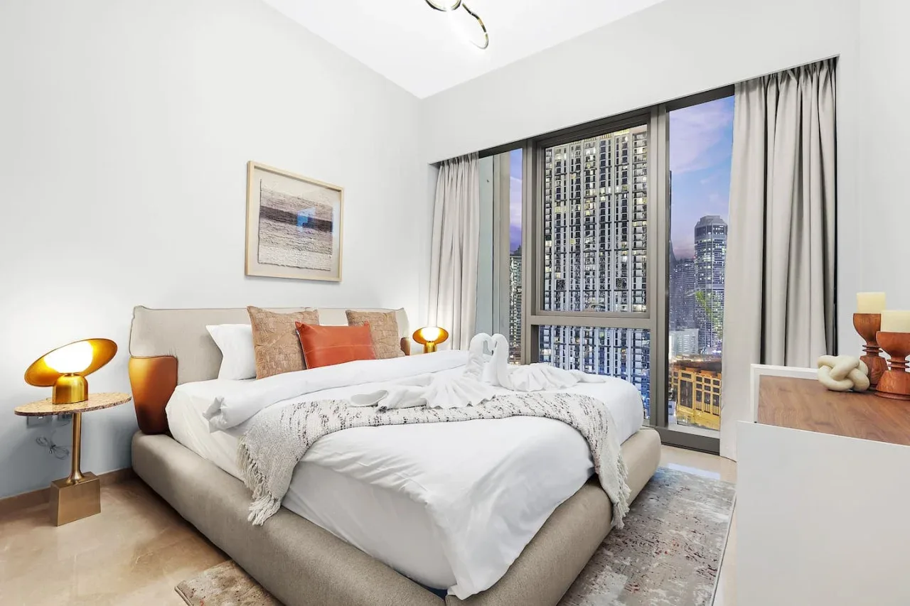 Un dormitorio con la cama tendida junto a ventanas altas con vistas a edificios de la ciudad.