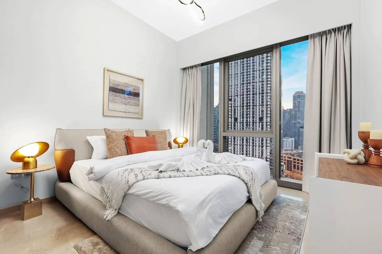 Un dormitorio con una cama tendida junto a grandes ventanas con vistas a edificios de la ciudad.