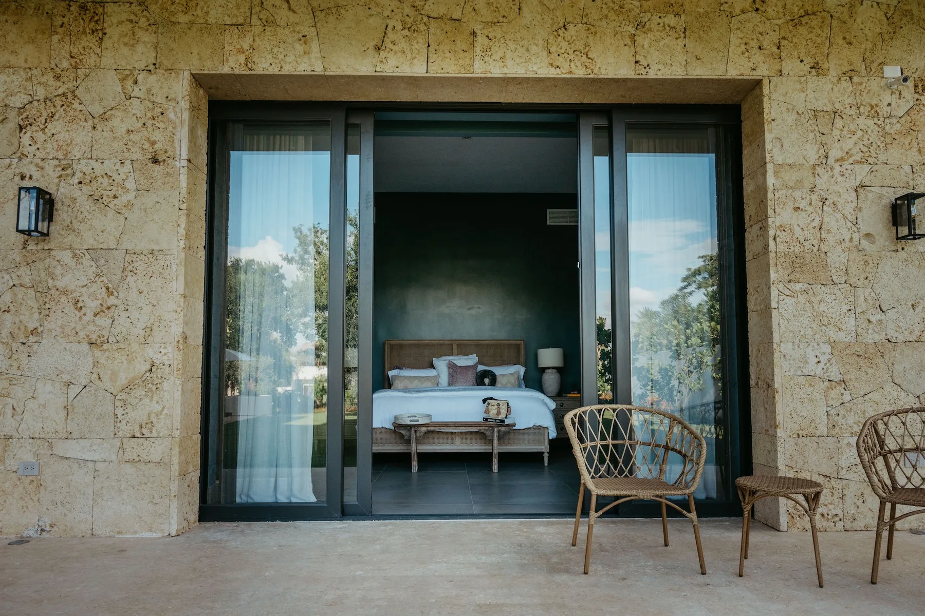 Un dormitorio da a un patio por amplias puertas de vidrio, con una cama visible en el interior.