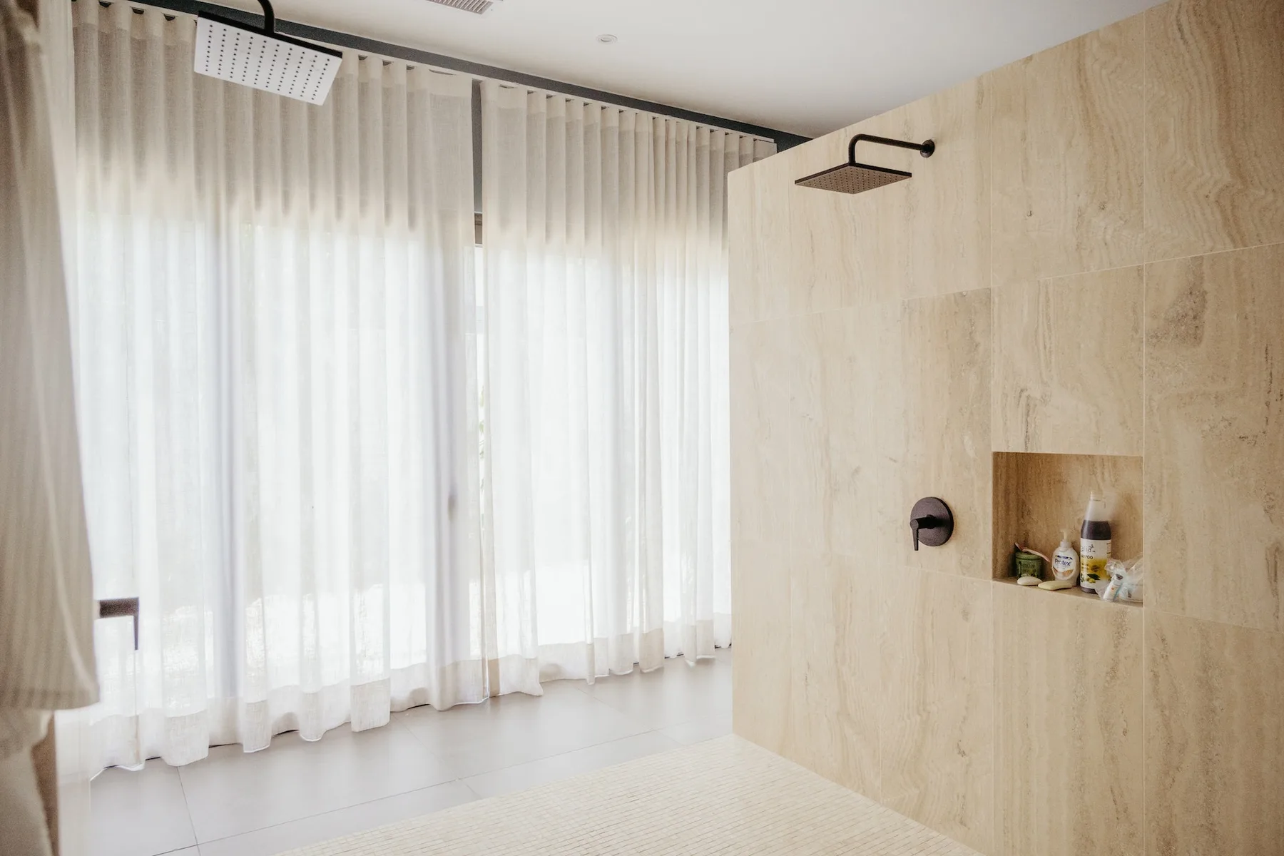 Una zona de ducha alicatada junto a una gran ventana con cortinas translúcidas.