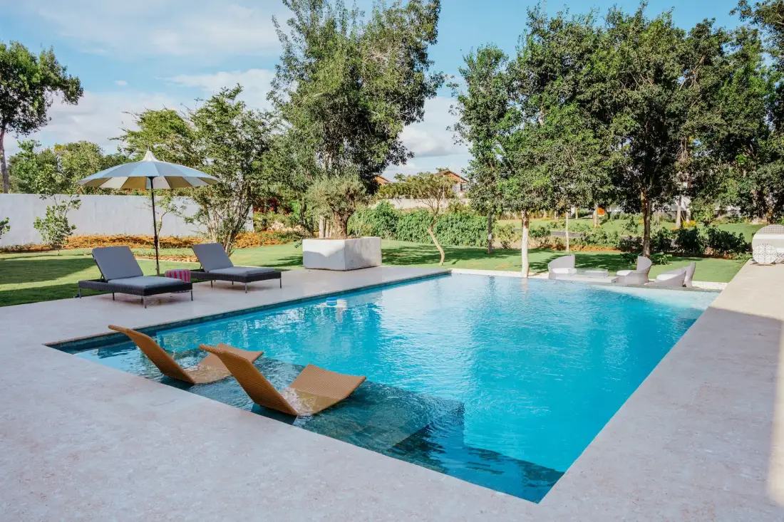 Una piscina al aire libre con tumbonas dentro del agua está junto a un jardín ajardinado.