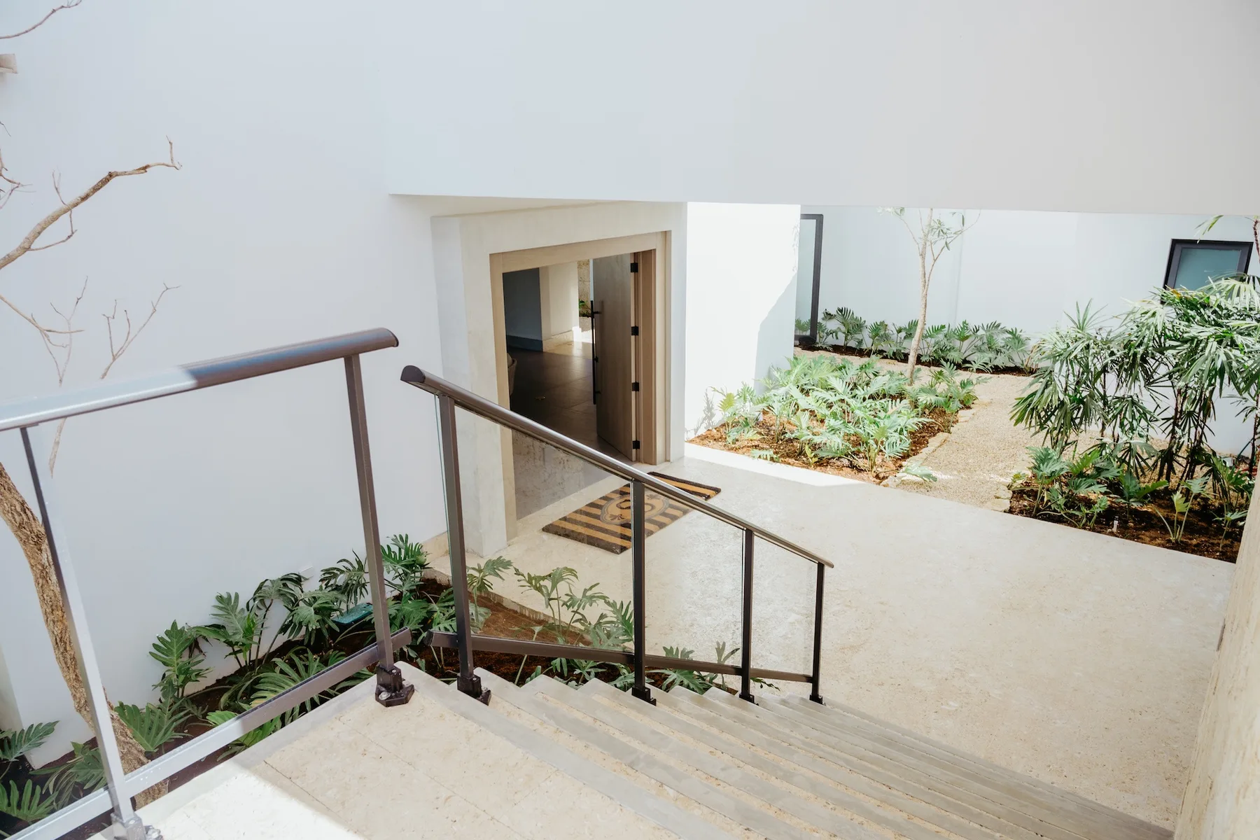 Una escalera exterior luminosa desciende junto a un patio ajardinado y una puerta abierta.