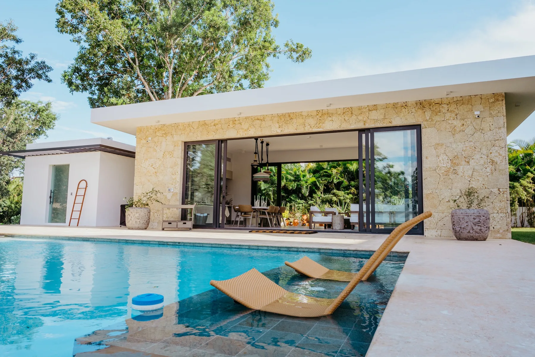 Una piscina junto a una casa moderna, con una zona de estar abierta y árboles a la vista.