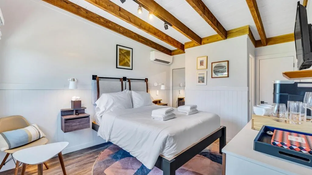 Un dormitorio luminoso con una cama tendida bajo vigas de madera expuestas.