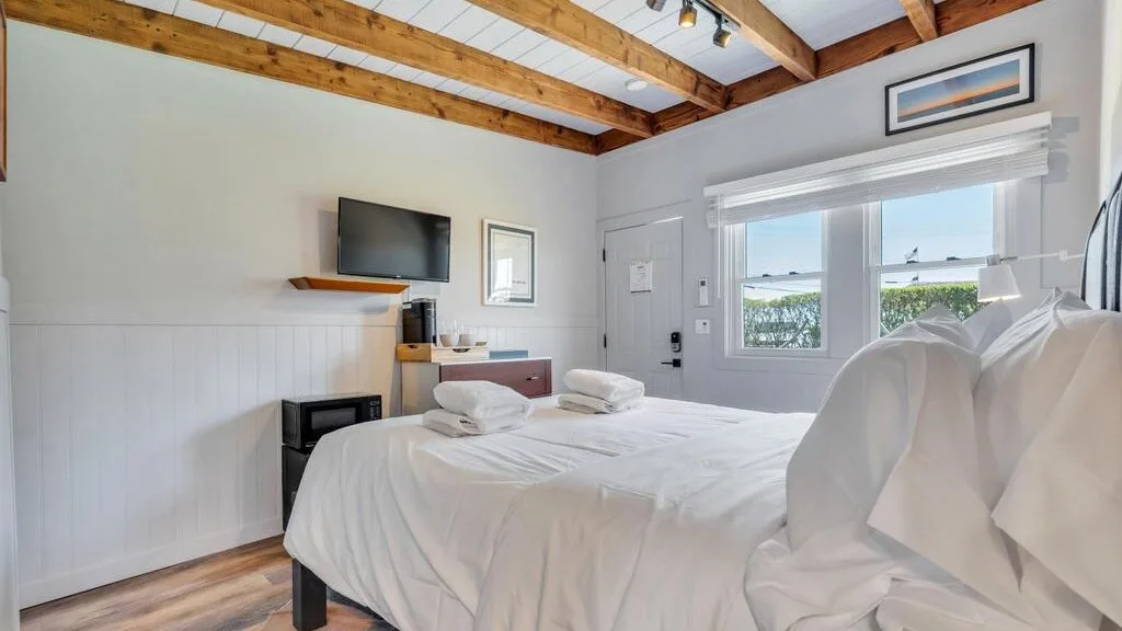 Un dormitorio luminoso con una cama bien tendida, un televisor en la pared y ventanas.