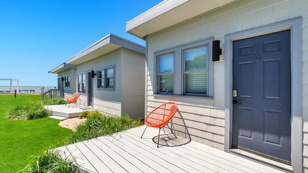 A row of small buildings opens onto a wooden deck beside a grassy lawn.