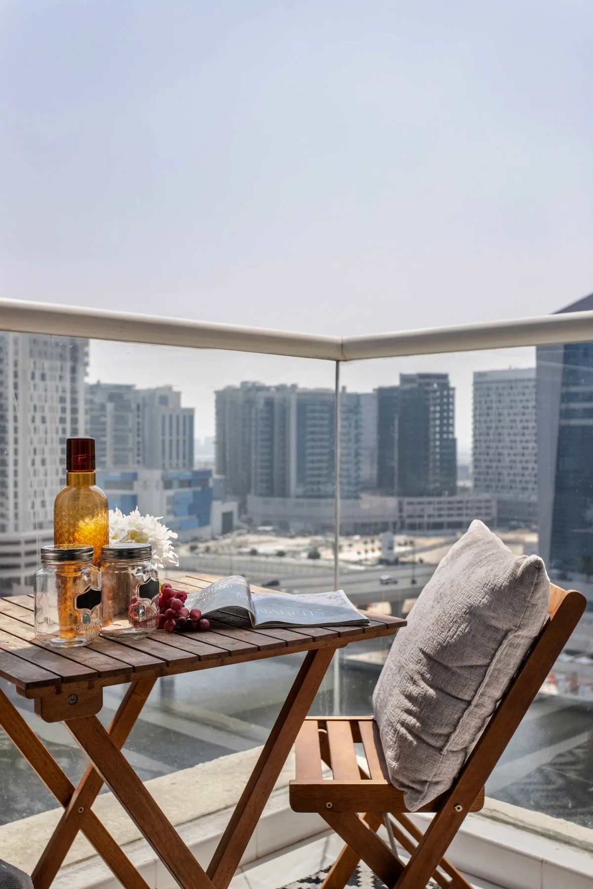 Una zona de estar en un balcón da a un horizonte urbano, con una mesa de madera y una silla con cojín.