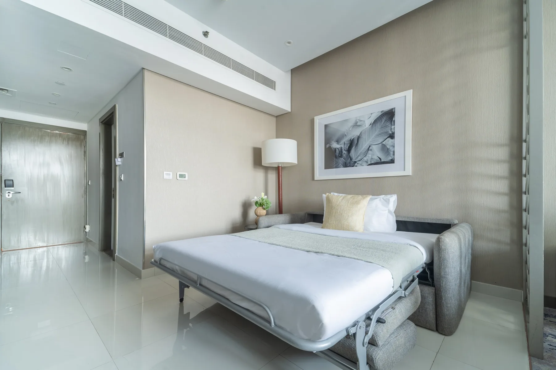 Un dormitorio con una cama, una obra enmarcada y una lámpara de pie.
