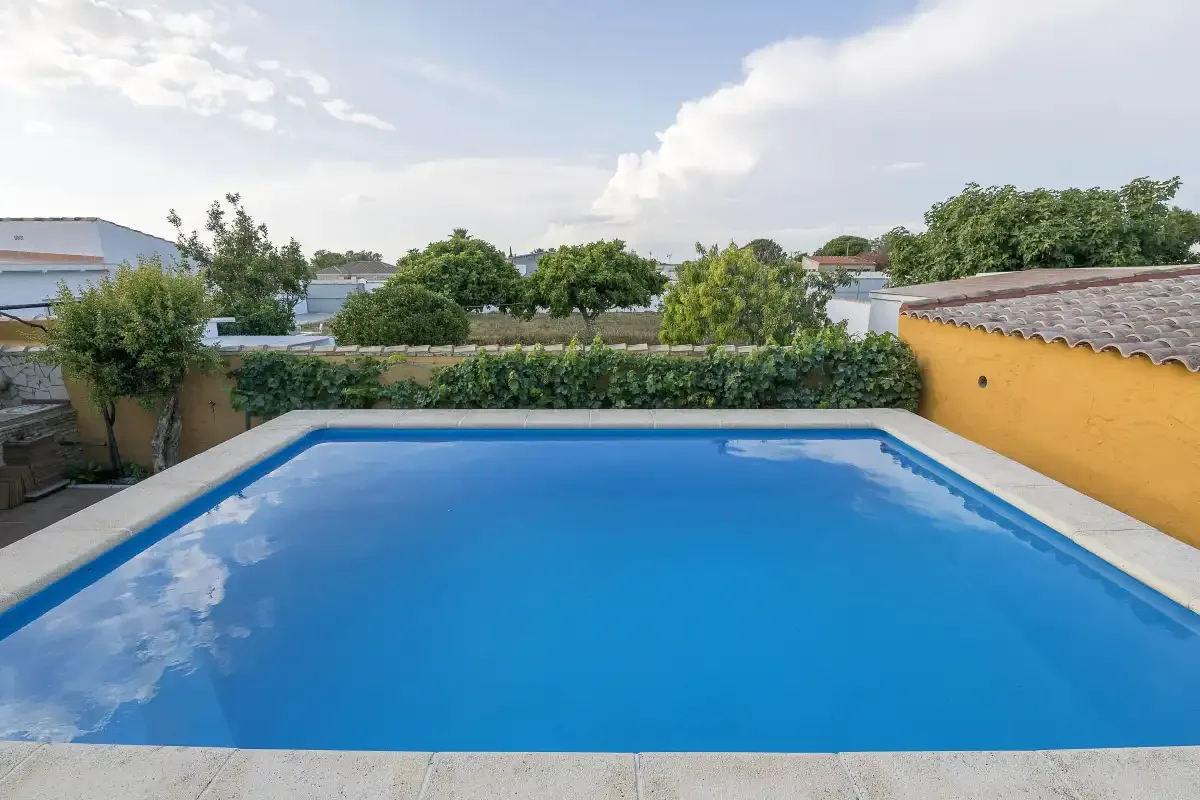 An outdoor swimming pool is bordered by paved decking, a yellow wall, and greenery.