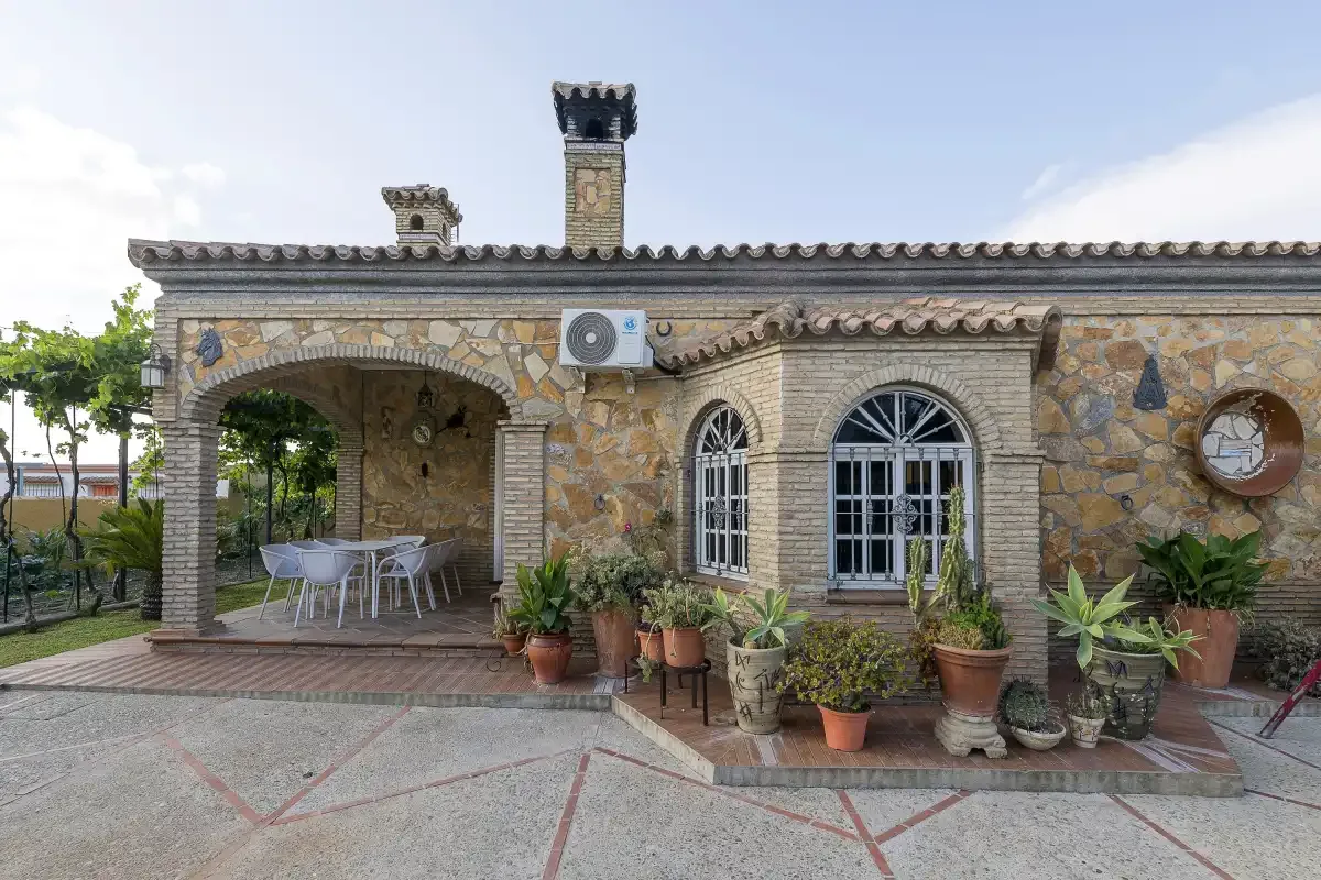 A stone house facade with an arched patio, barred windows, and potted plants.