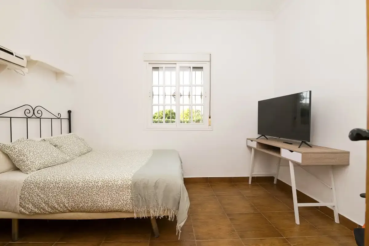 A bright bedroom with a bed, a window, and a television on a desk.