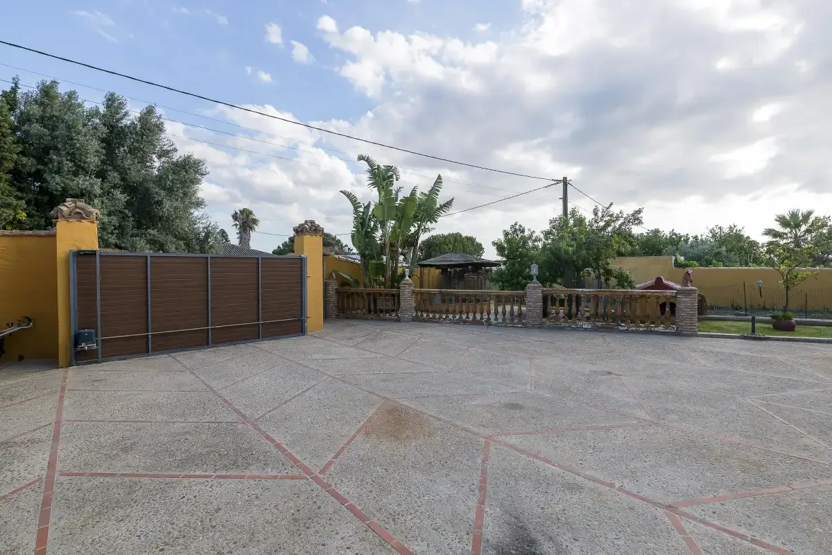 A paved outdoor courtyard with a wide gate and trees beyond.
