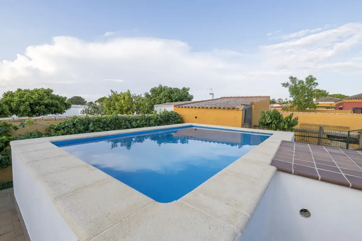 An outdoor swimming pool sits beside a paved patio under a partly cloudy sky.