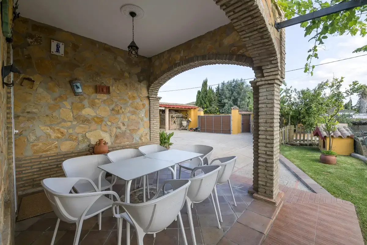 A covered patio with a dining table and white seating overlooks a garden and paved courtyard.