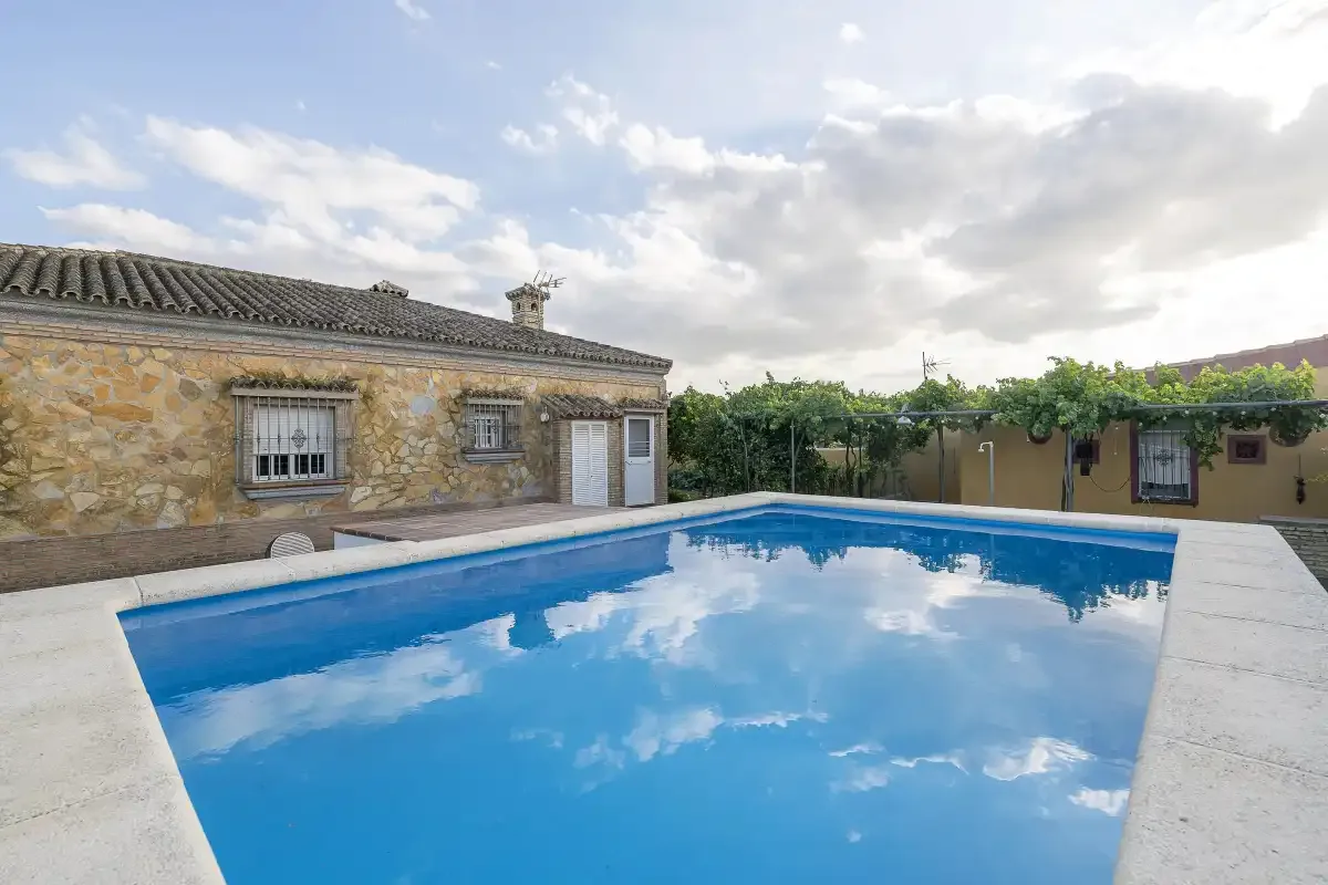 An outdoor swimming pool sits beside a stone house and a low garden wall.