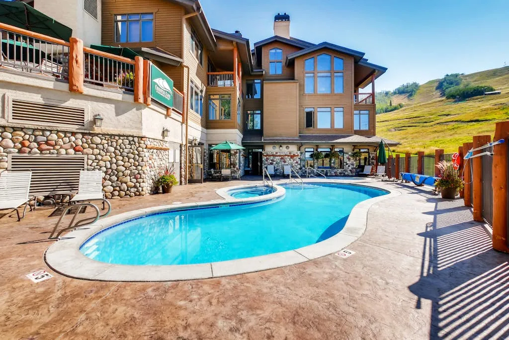 An outdoor swimming pool sits beside a multi-story building, with hills in the background.