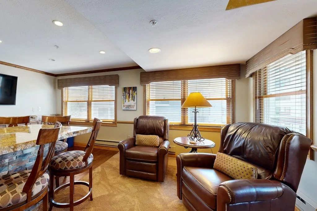 A furnished living room with leather seating, a dining table, and large windows.