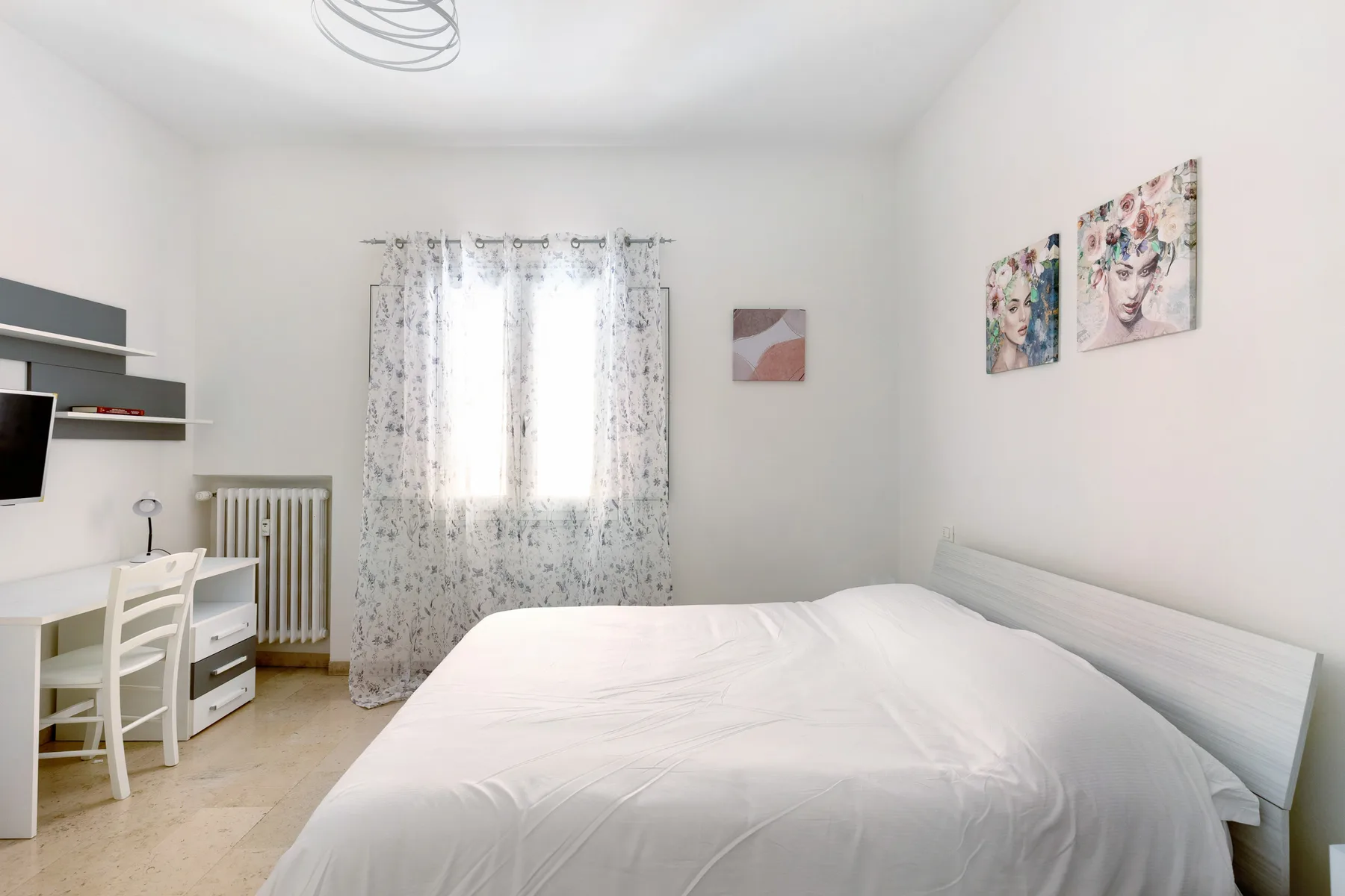 A bright bedroom with a white bed, a desk, and a window with patterned curtains.