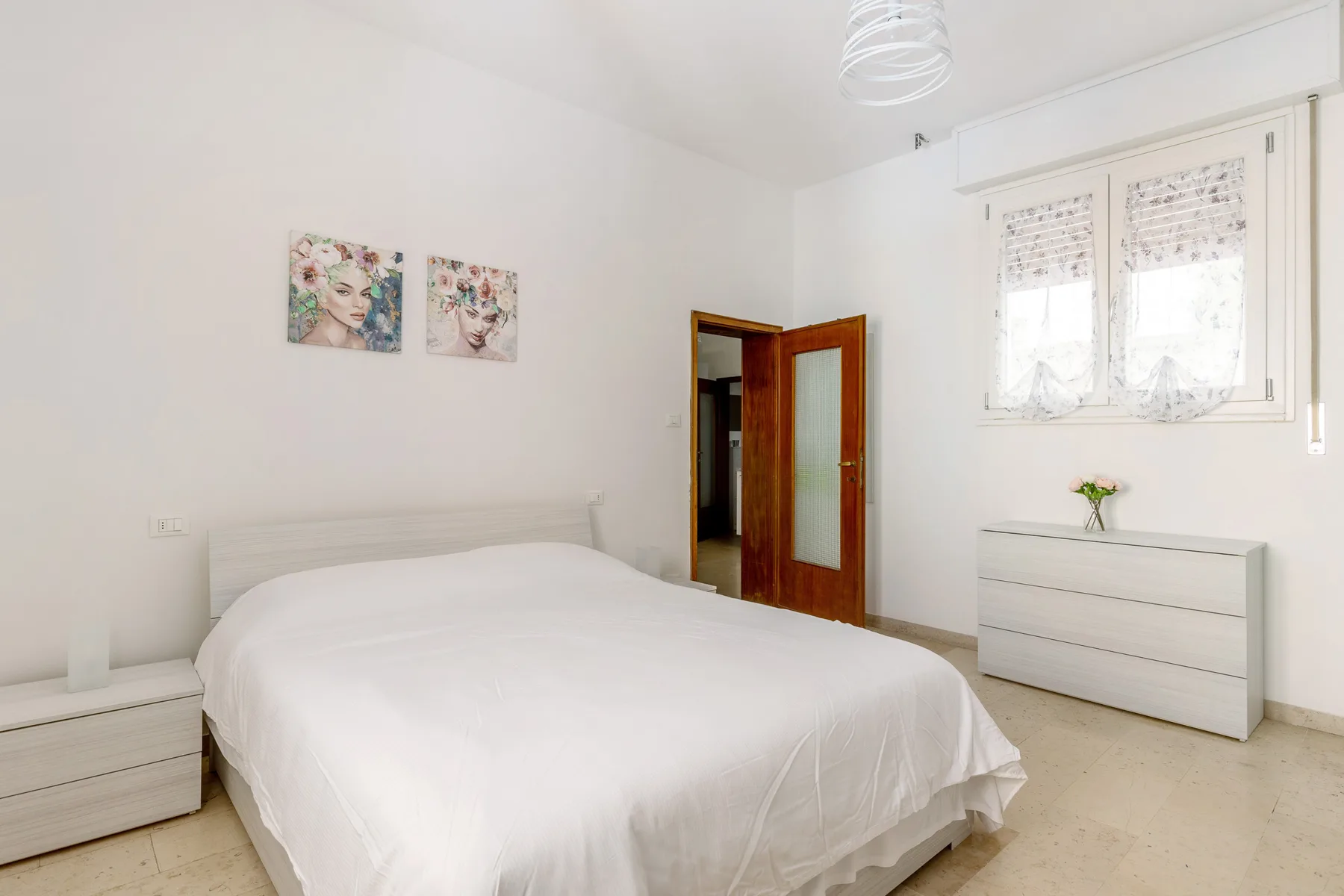 A bright bedroom with a white bed, framed artwork, and a window.