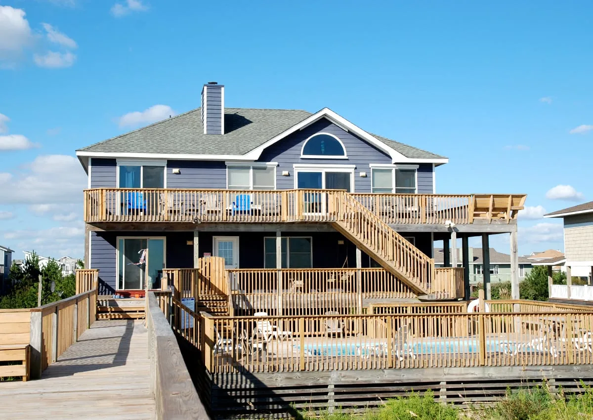 A large house with a raised wooden deck, stairs, and a fenced swimming pool.