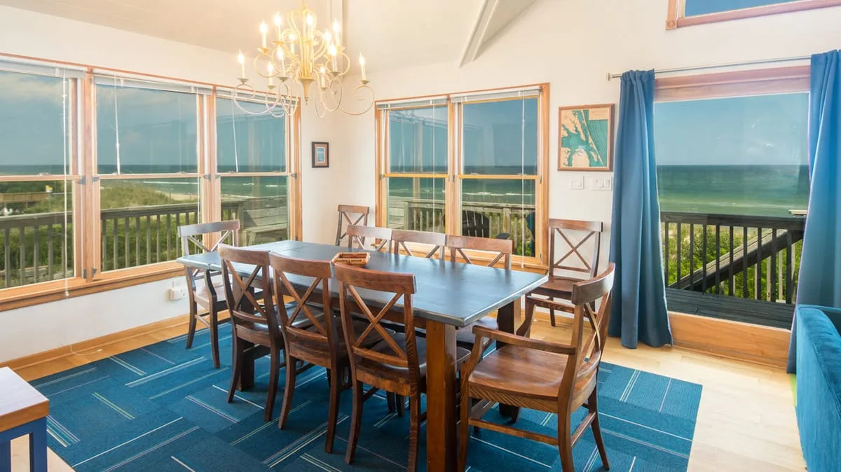 A dining area with a long wooden table beside large windows overlooking a green landscape.