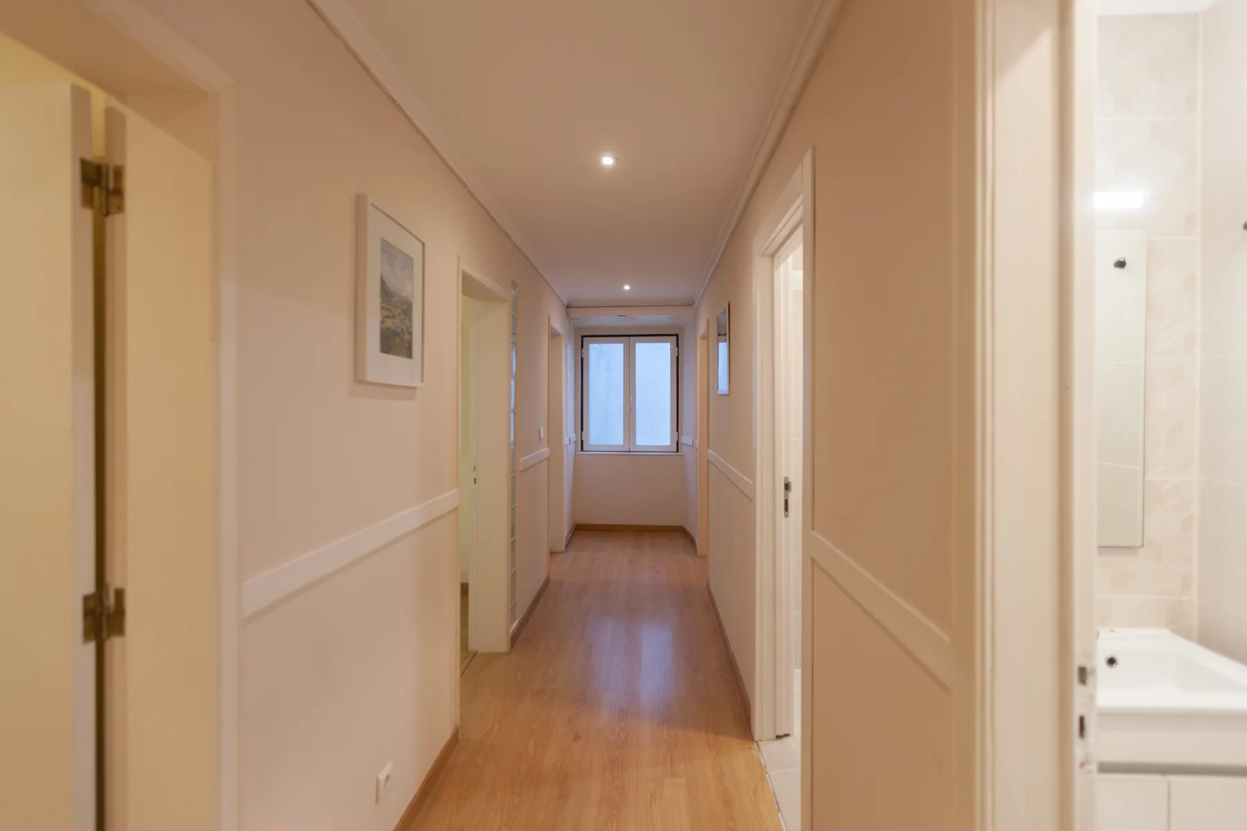 A narrow hallway with wood flooring leads to a window at the far end.
