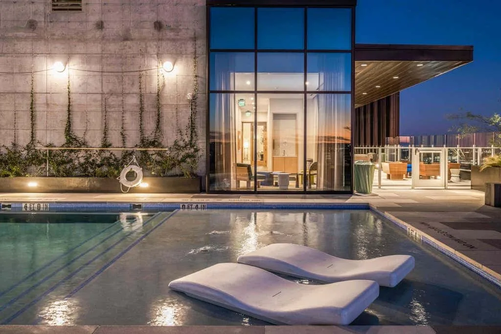Una piscina exterior junto a un edificio moderno, con asientos de descanso dentro del agua.