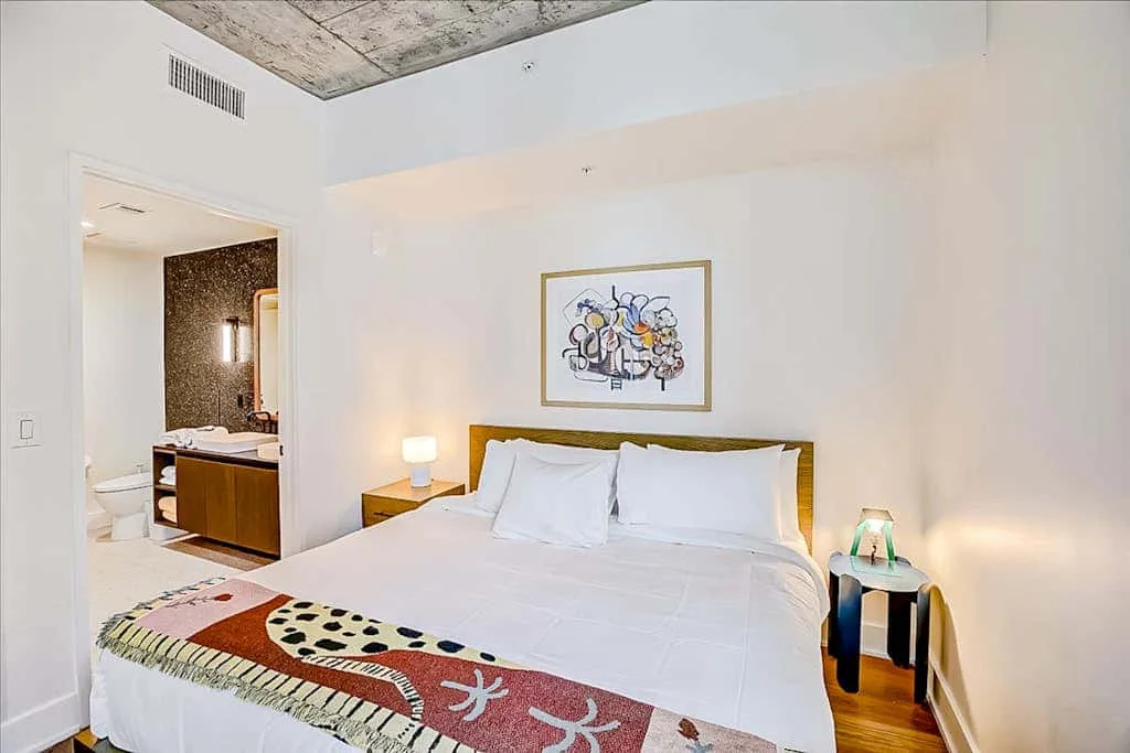 Un dormitorio luminoso con una cama tendida y una obra enmarcada encima.
