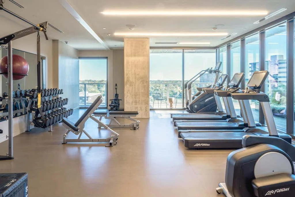 Un gimnasio con máquinas de ejercicio y estantes de pesas junto a grandes ventanas.
