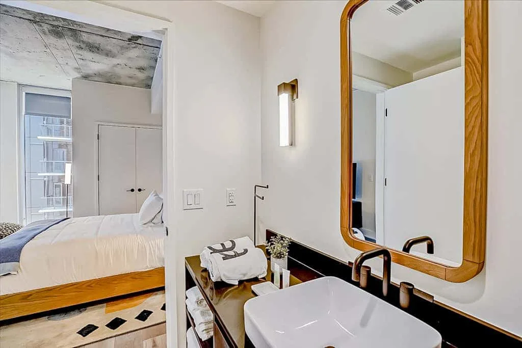 Un tocador de baño con un espejo amplio, junto a un dormitorio con la cama tendida.