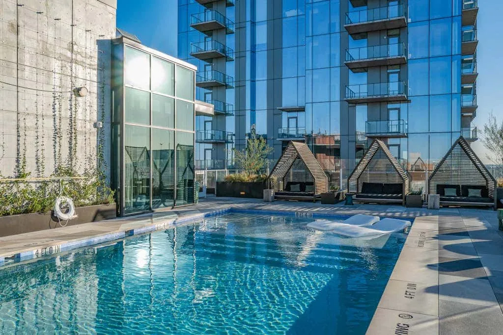 Una piscina en la azotea está junto a un edificio acristalado y pequeños refugios con tejados puntiagudos.