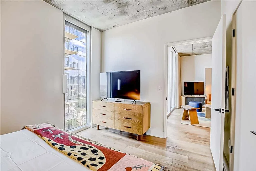 Un dormitorio luminoso con un televisor sobre una cómoda baja de madera junto a una ventana grande.