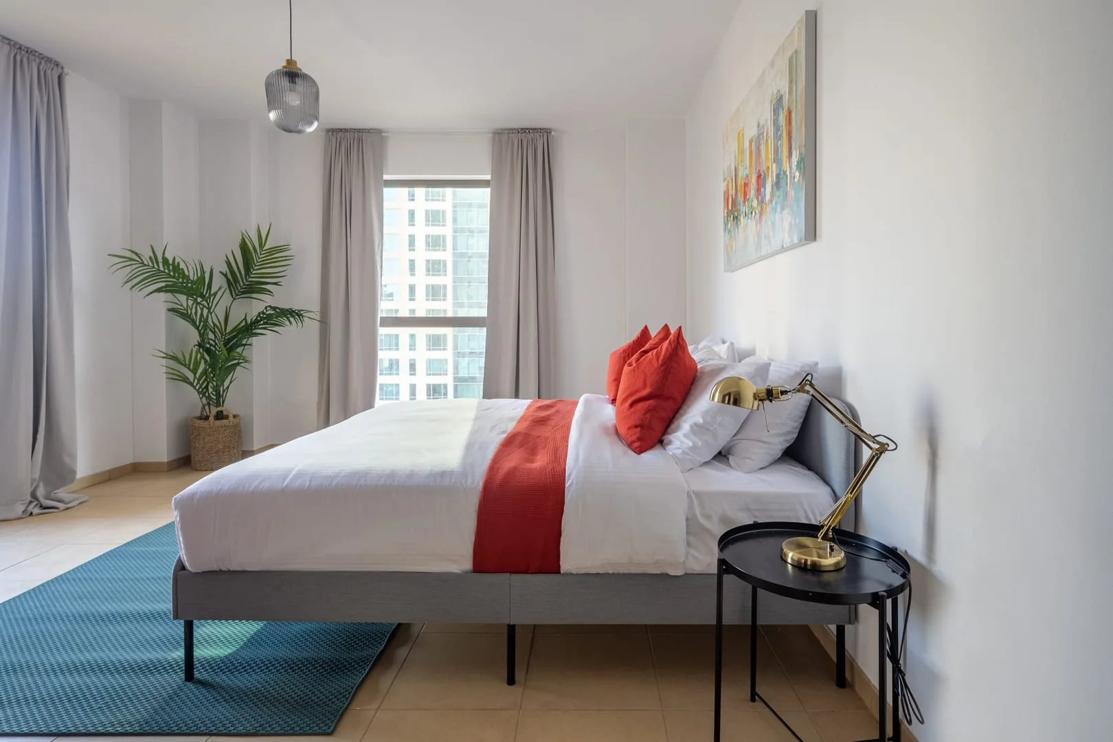 Un dormitorio luminoso cuenta con una cama con detalles rojos y una planta junto a la ventana.