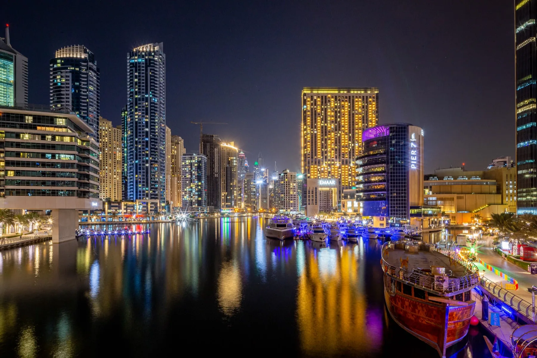 Un paseo marítimo urbano de noche, con edificios iluminados reflejados en el agua.
