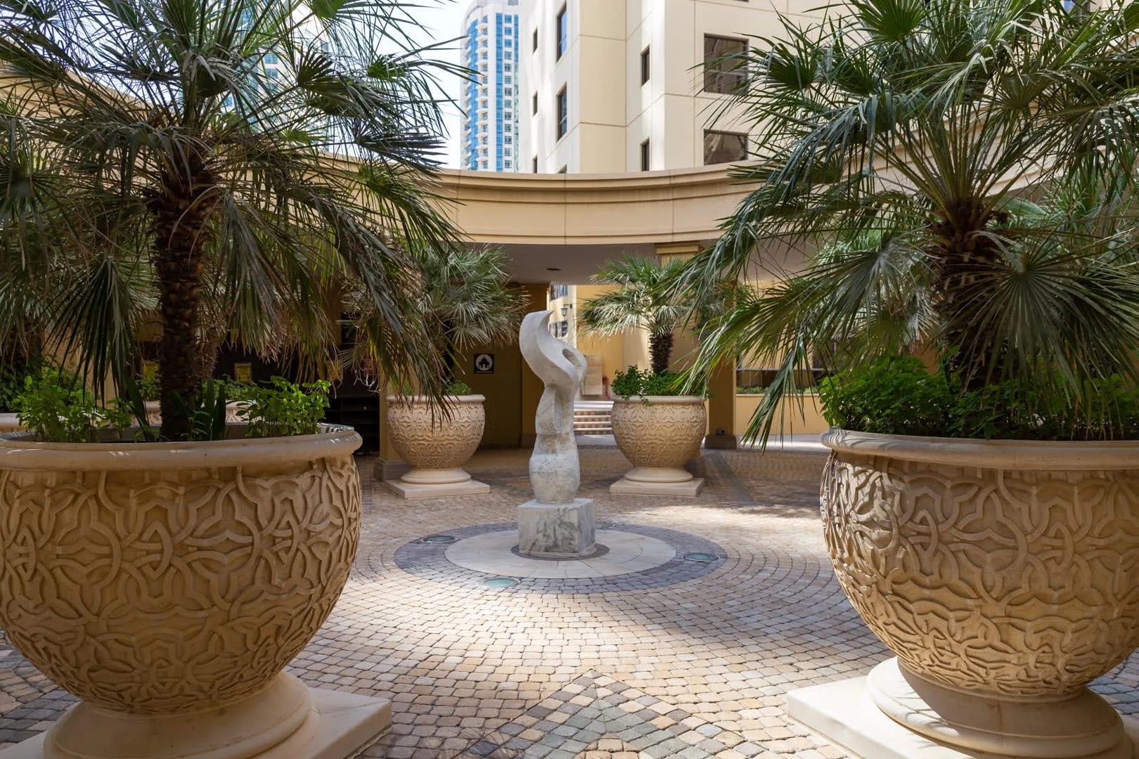Un patio exterior presenta una escultura central, pavimento estampado y frondosas palmeras.
