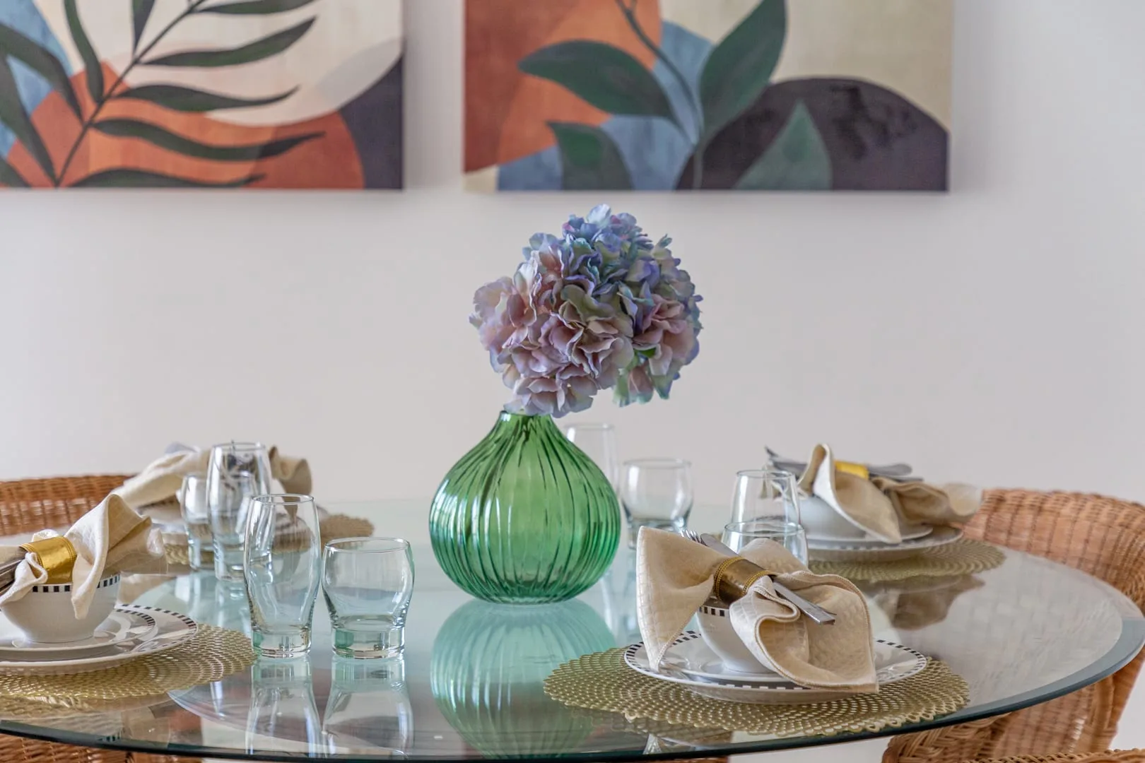 Una mesa de comedor está puesta con cristalería y un jarrón de flores bajo cuadros botánicos.