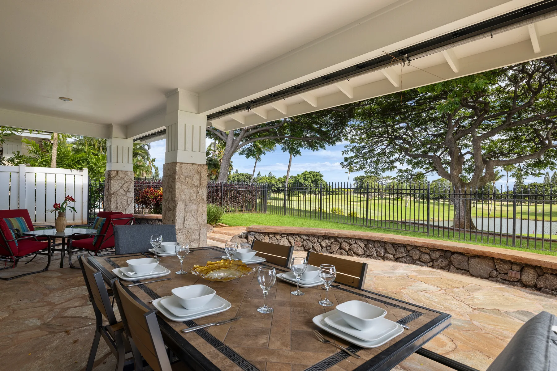 Comedor en un patio cubierto, con la mesa puesta frente a un jardín cercado y árboles.