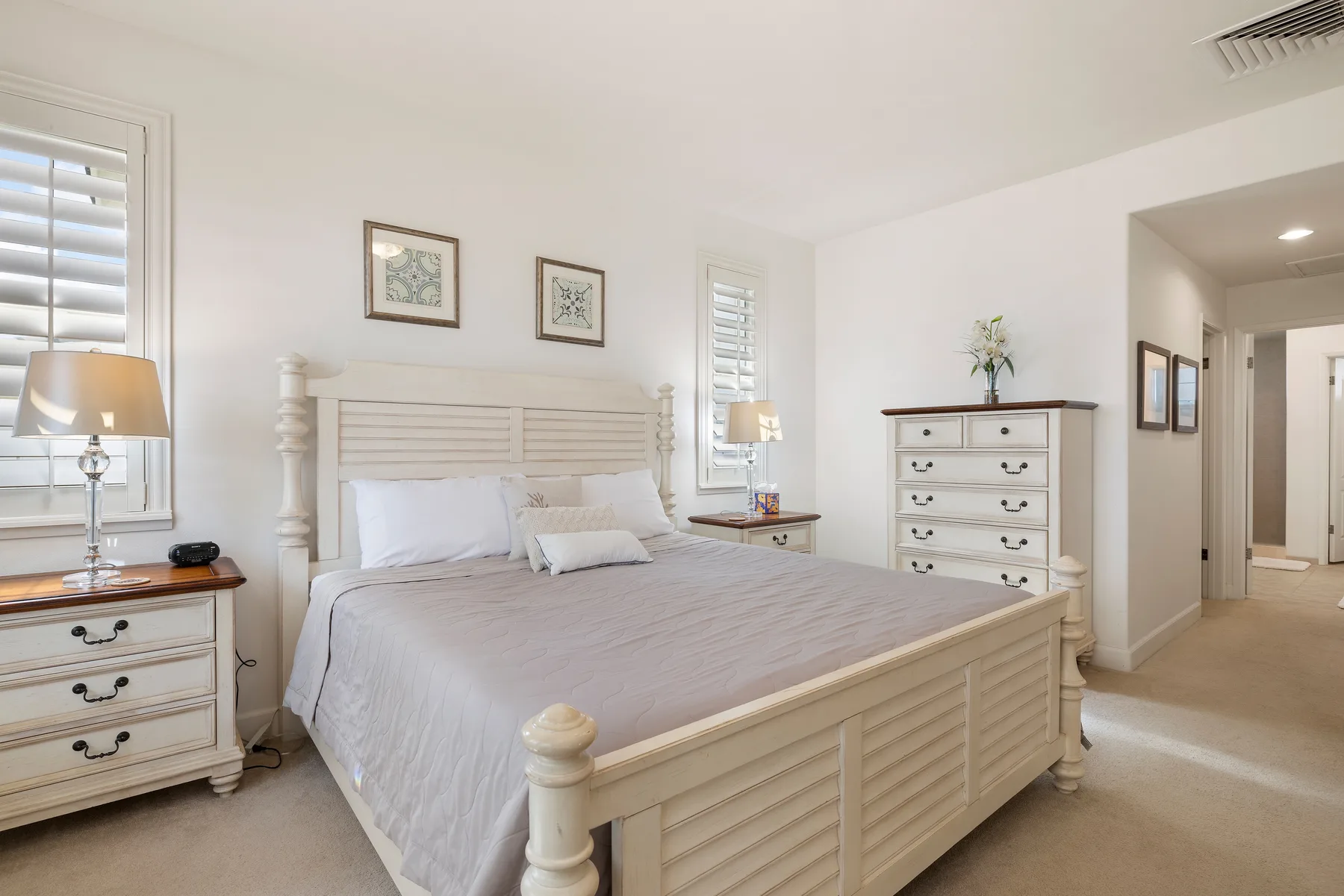 Un dormitorio luminoso con una cama grande, muebles blancos y ventanas con persianas.