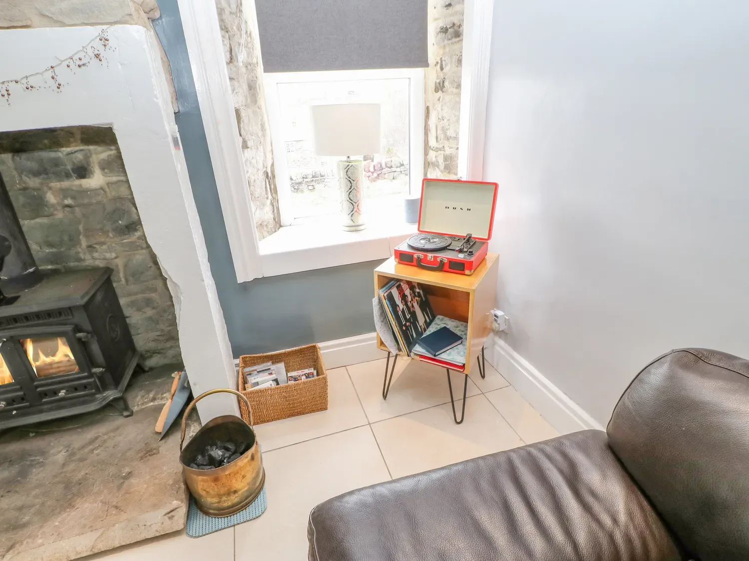 Un rincón con una chimenea encendida, un cubo de latón para carbón, una cesta de mimbre, un mueble de madera para tocadiscos con un tocadiscos rojo y discos de vinilo, y un sillón de cuero en 1 Braeside, en Cowshill, cerca de Wearhead