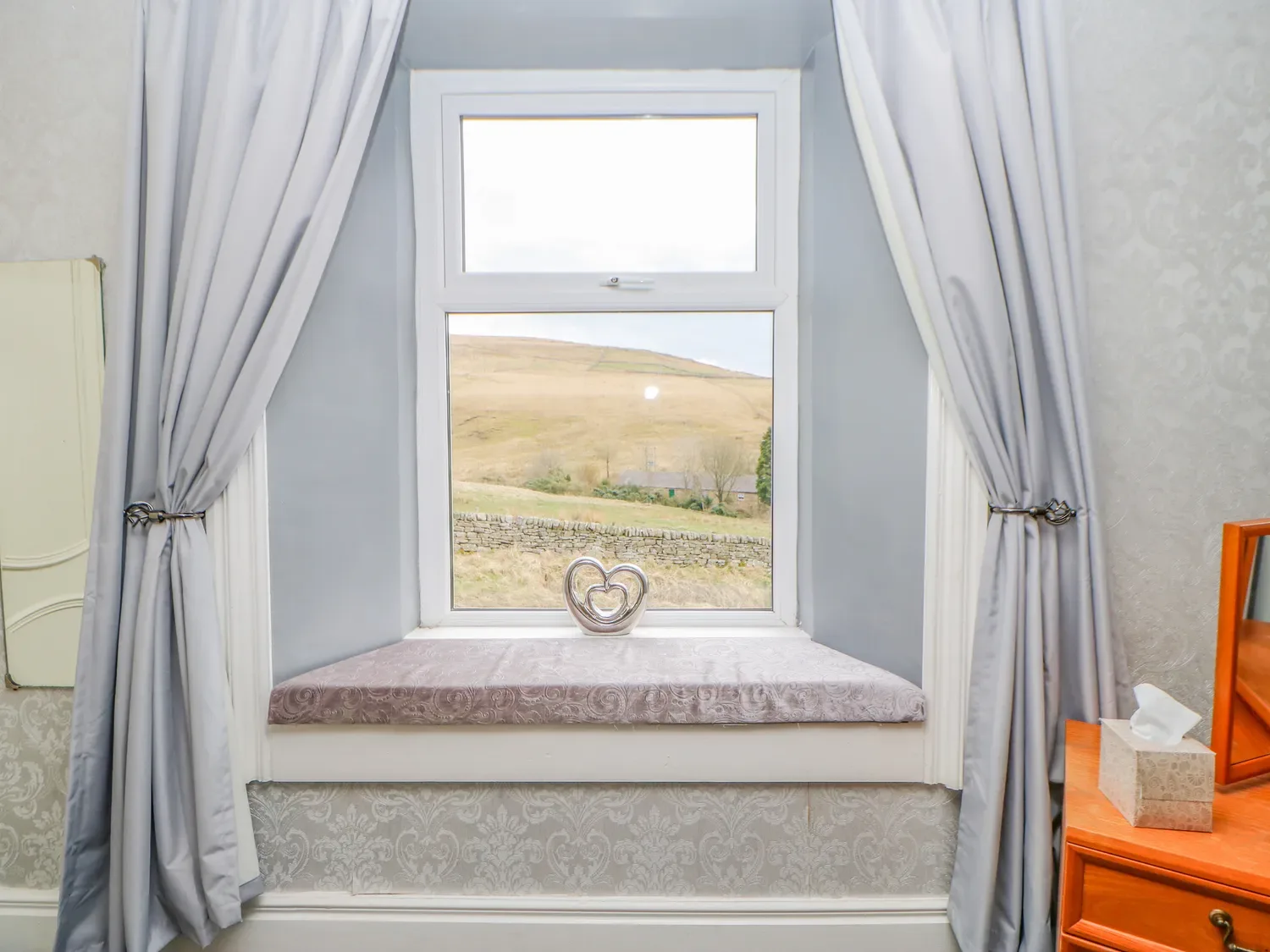 Una ventana con cortinas tiene un alféizar acolchado y un adorno en forma de corazón.