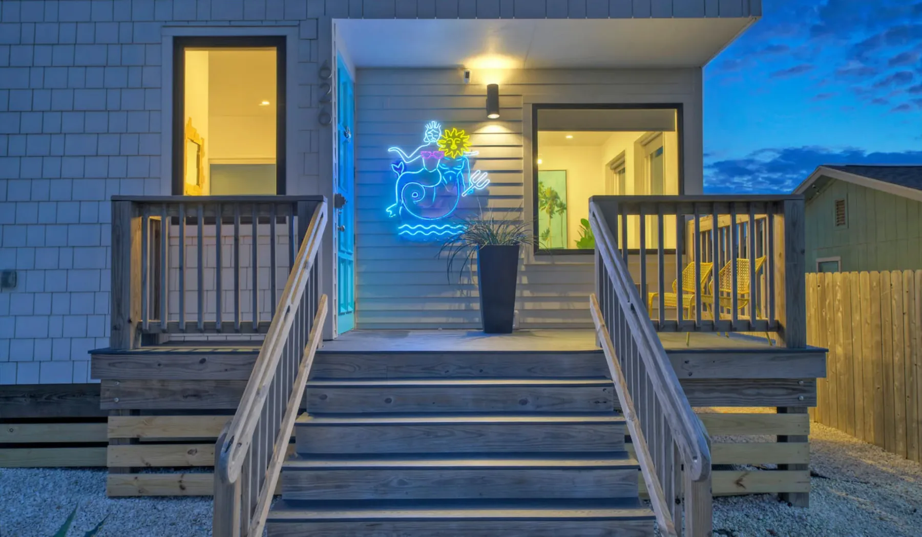 A wooden entrance deck with stairs leads to a brightly lit house at dusk.