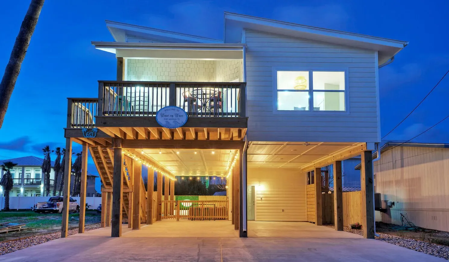 A raised coastal-style house stands on supports, with a balcony above the driveway.