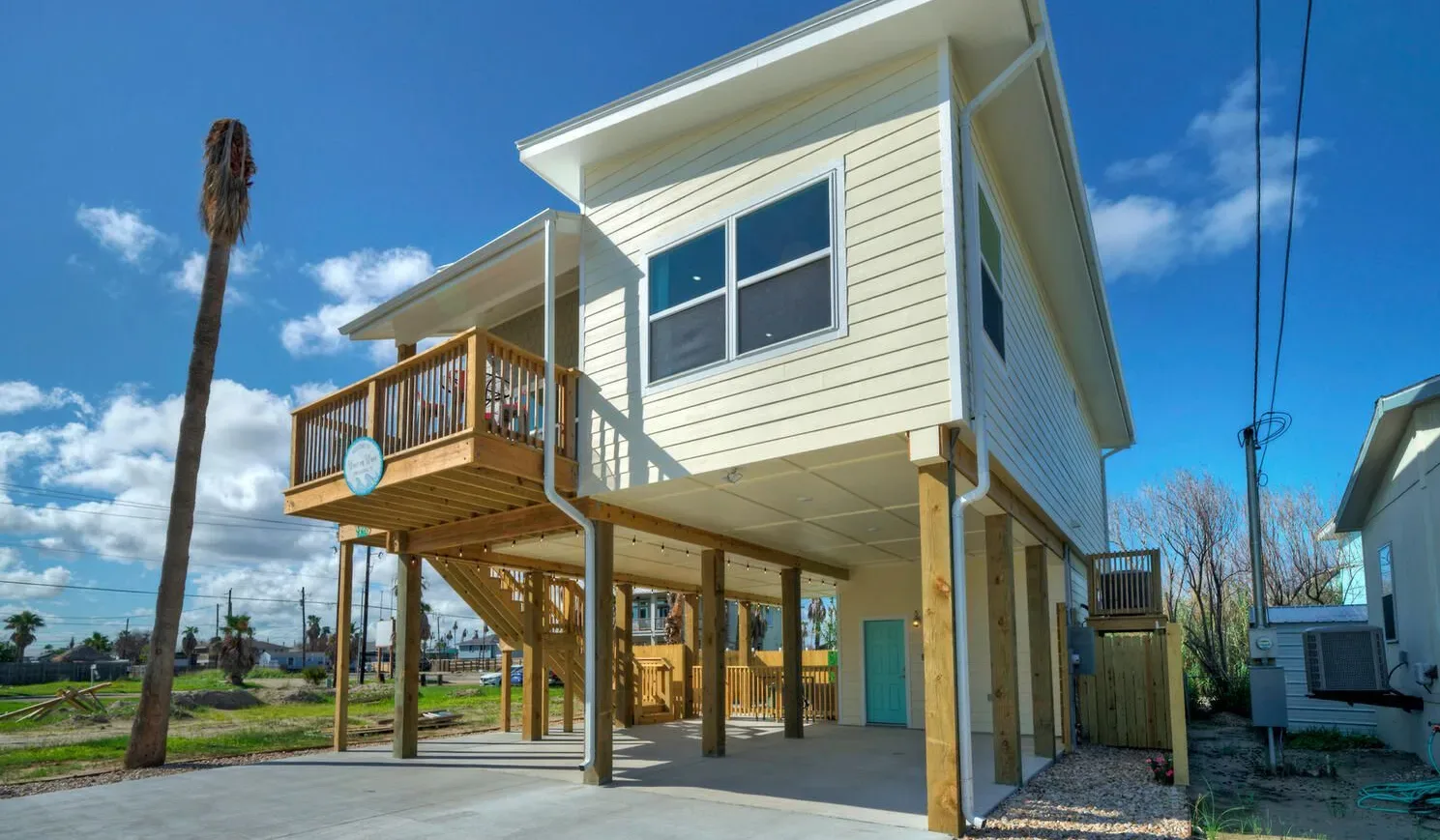 A raised light-colored house stands beside a wooden deck and open ground-level space.